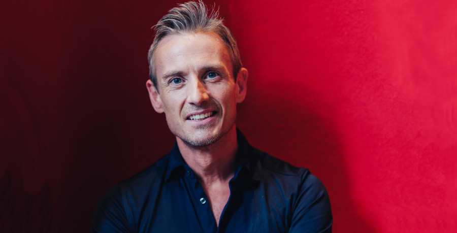 Conductor Alexander Shelley is wearing a black shirt and standing against a red background. He is looking straight at the camera and smiling. 