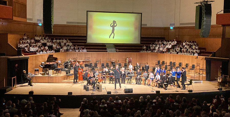 An orchestra plays on a wooden panelled stage, with a large screen above them showing a  yellow background and a stick-like human figure