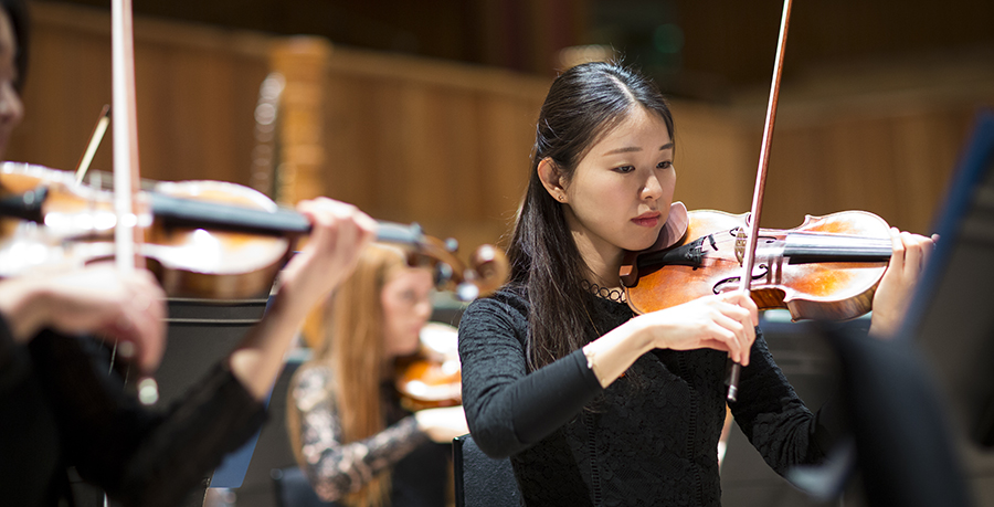 An image of a female violinist holding her instrument with a wooden panelled stage in view behind