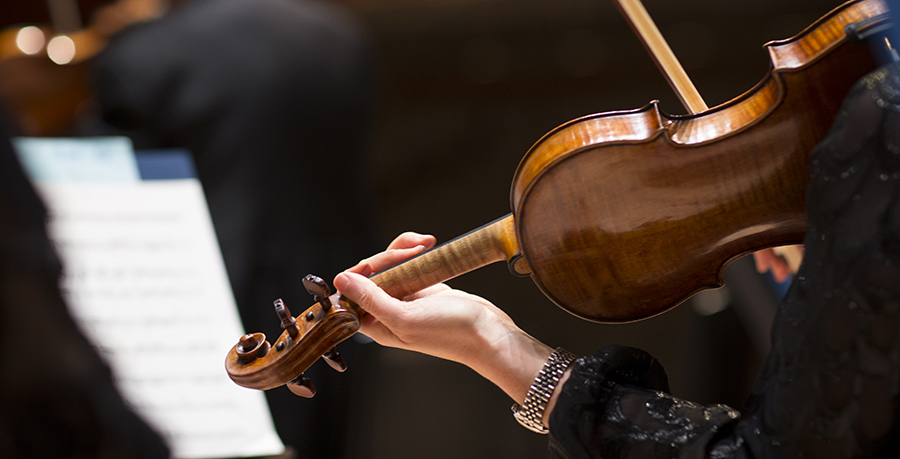 An image of a hand holding a violin, a watch is visible on their wrist and sheet music is visible in the background