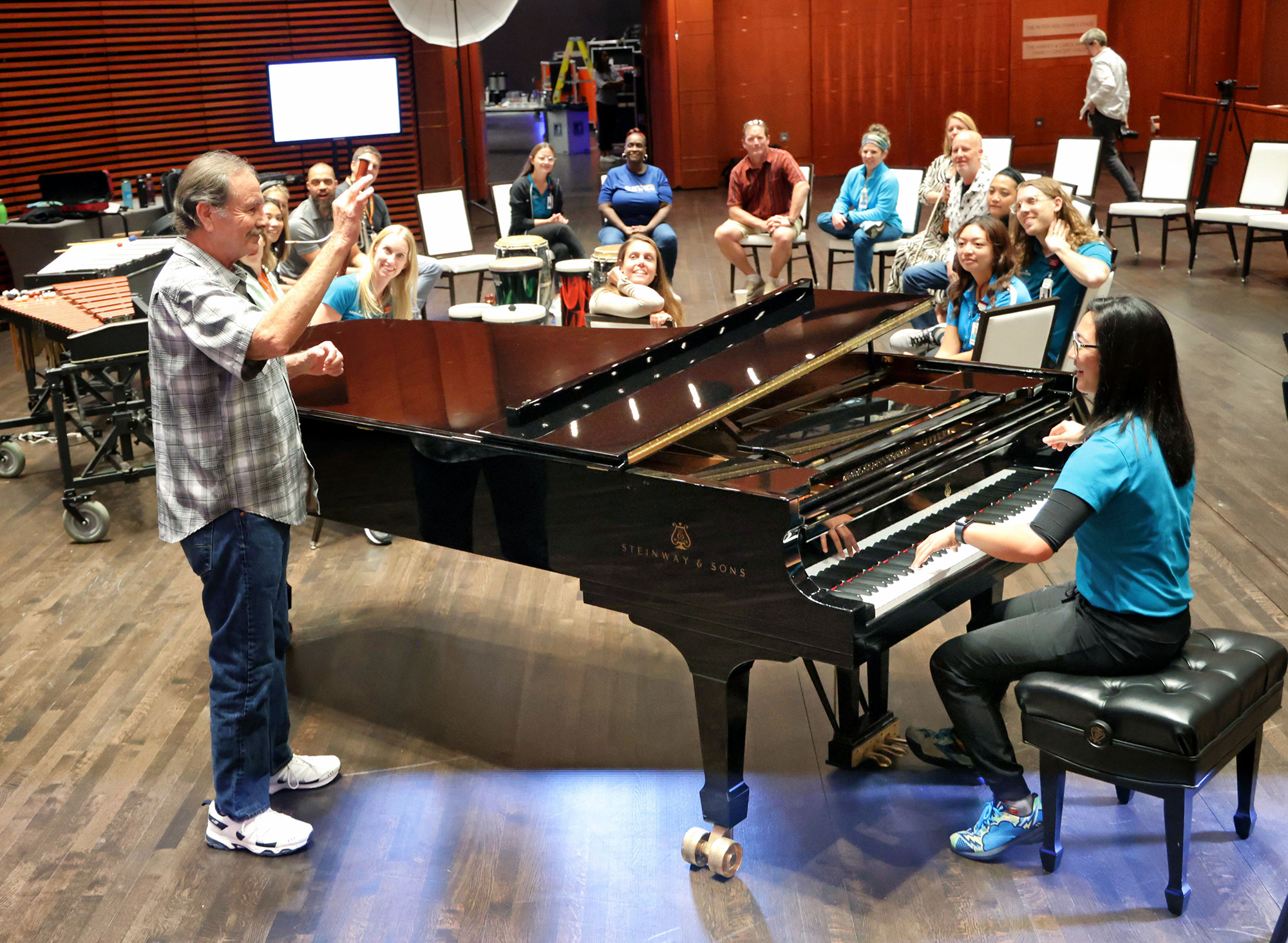 An image of a pianist playing a grand piano with a group of people sitting around it. There is a man conducting the group.