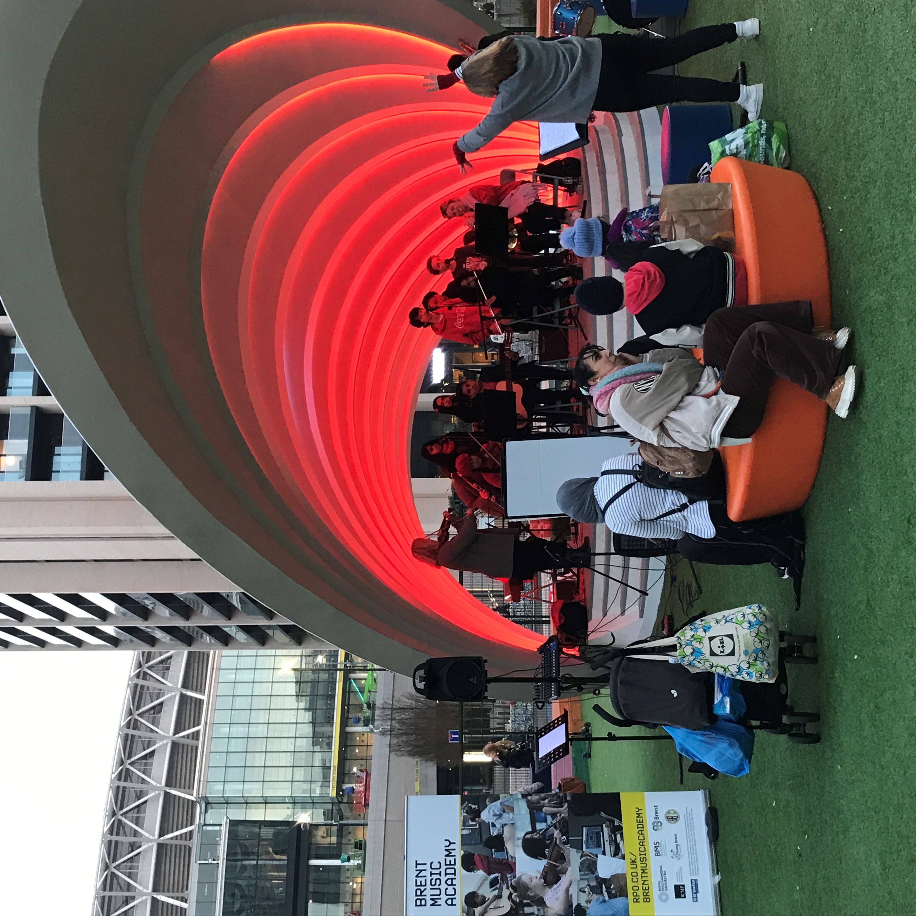 RPO players and young musicians playing in a large shell-like structure in with red lighting in Wembley Park