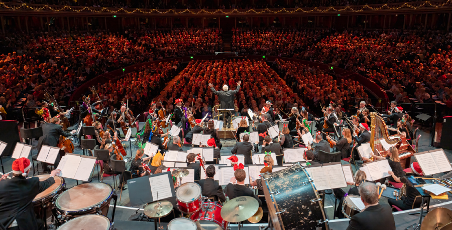 John Rutter conducting the audience form the podium at the Royal Albert Hall, with the Orchestra wearing Christmas accessories