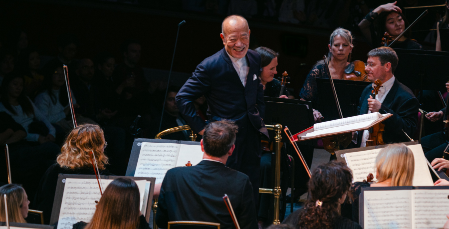 Joe Hisaishi smiling widely on the podium of the Royal Albert Hall as he looks at the second violins