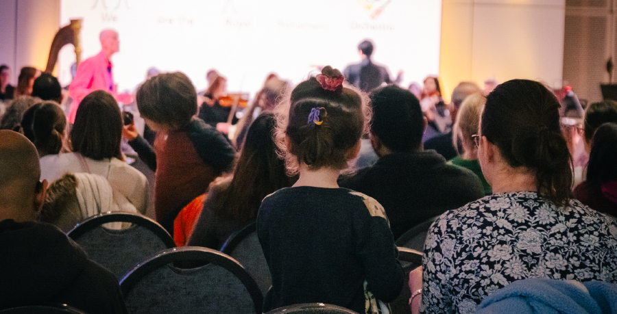 An audience of children and adults watching the RPO perform in Brent Civic Centre, with a presenter in a pink suit standing in front of the Orchestra