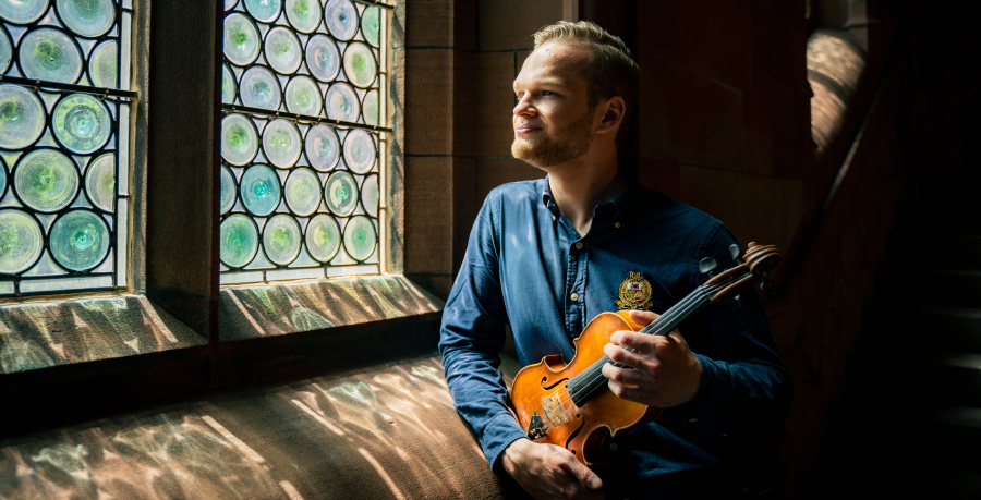 Savva Zverev in a blue shirt holding his violin and looking ut of a stained glass window